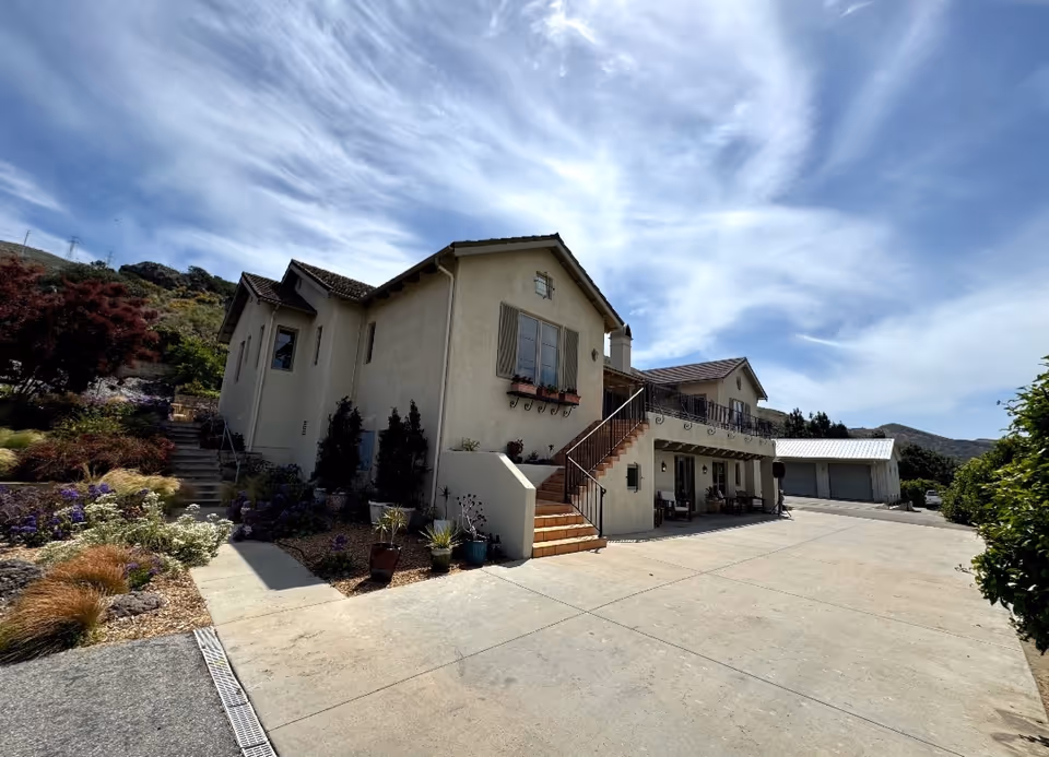 Exterior view of a two-story beige building with a tiled roof, stairs leading to a balcony, and a driveway. The building is surrounded by landscaped plants and trees under a partly cloudy sky.