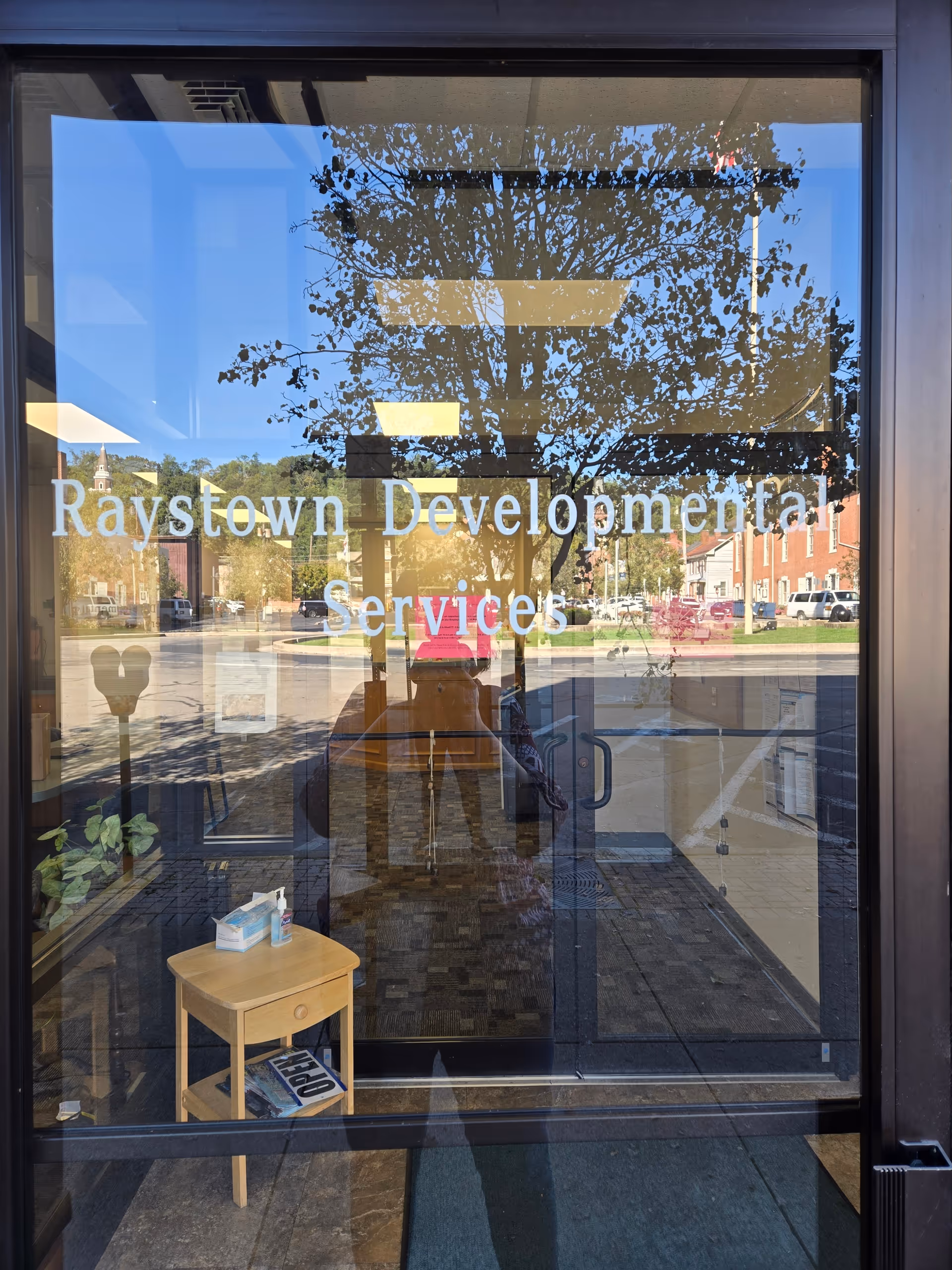 Glass entrance door with Raystown Developmental Services sign, reflecting trees outside and a small wooden table.