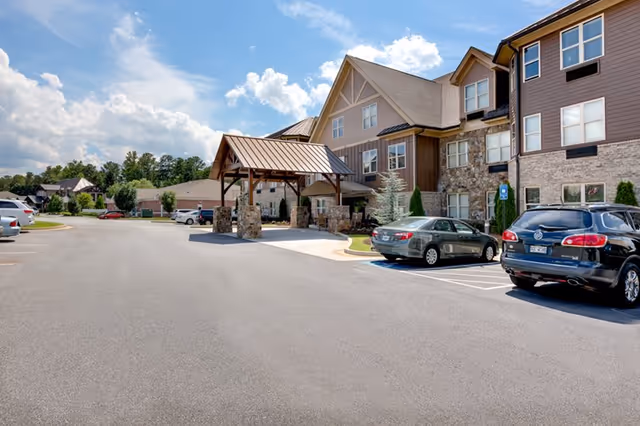 Front exterior of a senior living building with a covered porte-cochere, parked cars, and a driveway under a partly cloudy sky.