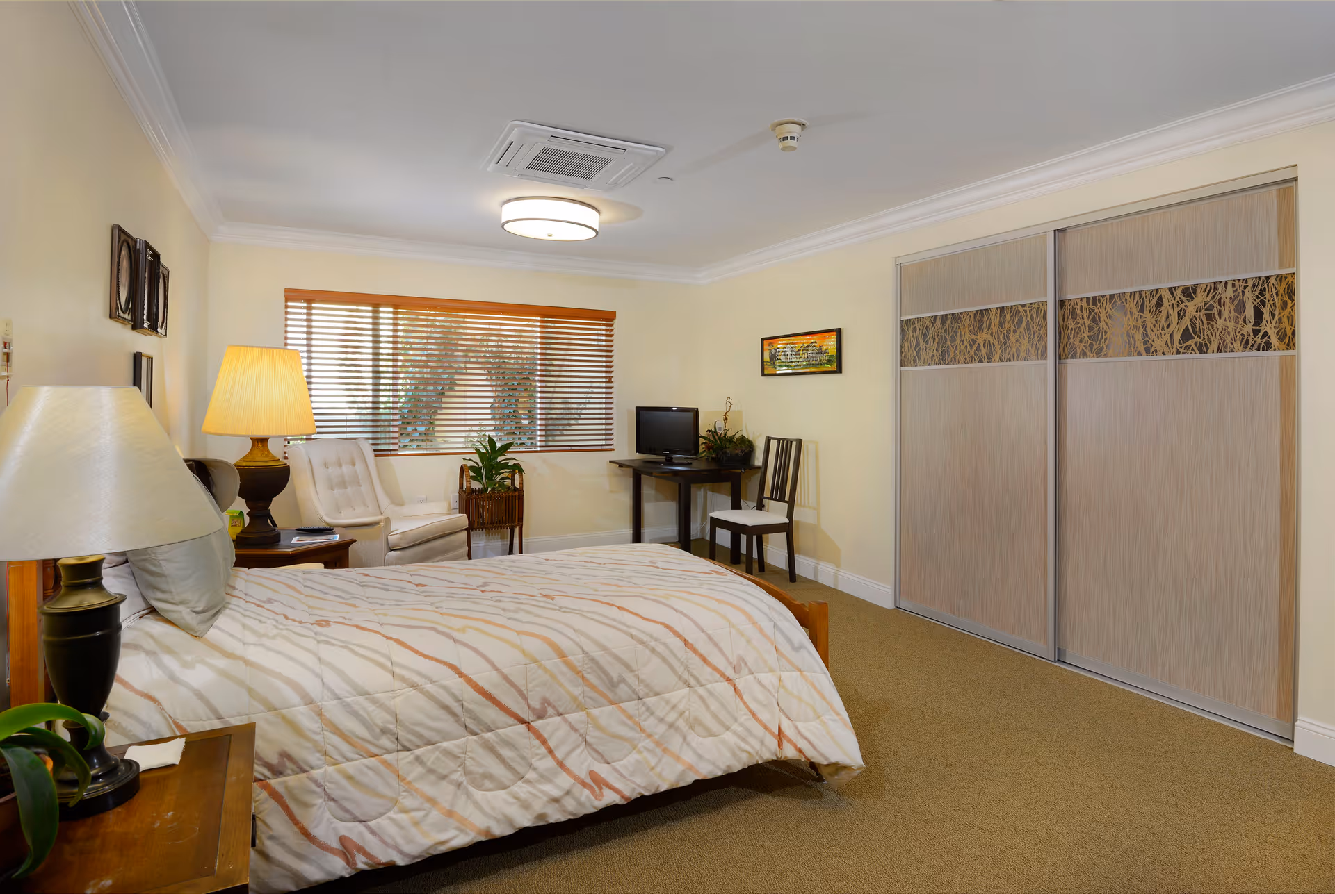A well-lit bedroom with a bed covered in a patterned comforter, two bedside tables with lamps, a white armchair near a window with wooden blinds, a small desk with a chair and a computer monitor, and a large closet with sliding doors featuring a decorative panel.