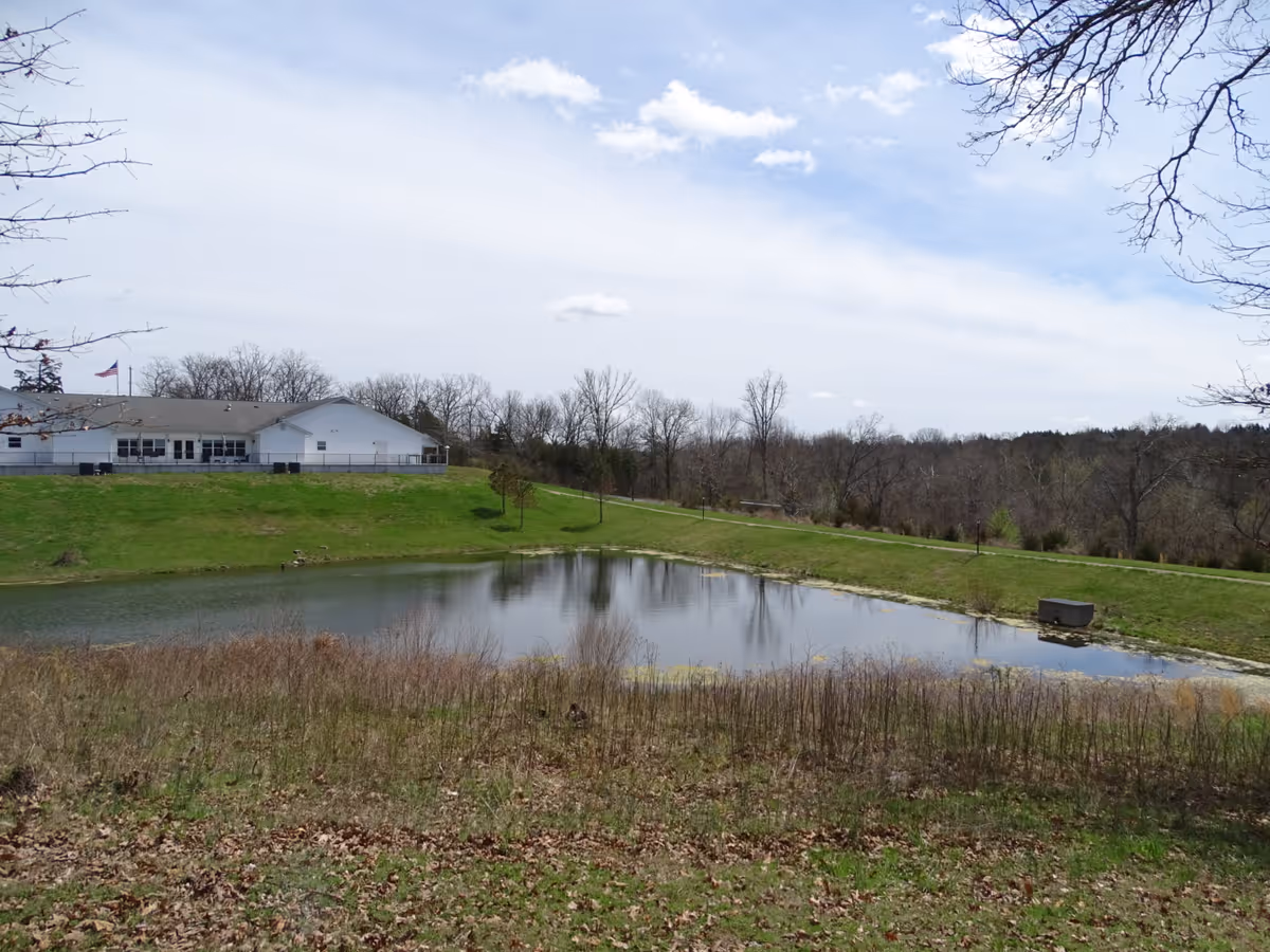 Outdoors scene with a pond, grassy slope, and the Pony Bird Home building in distance.