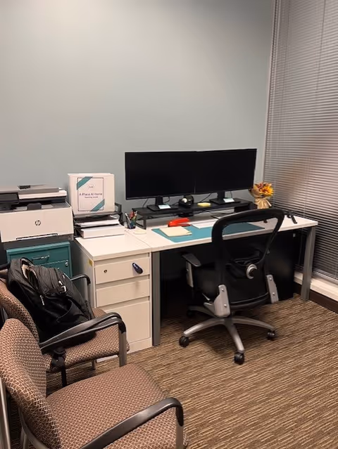 A tidy office desk with dual monitors, printer, and chairs in a small workspace.