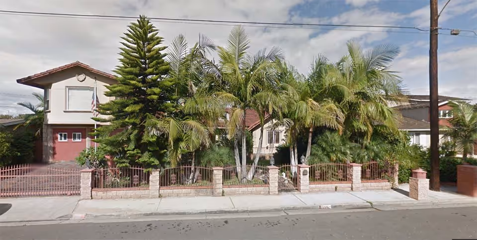 Front view of Newport Heights Manor with palm trees, a pink brick fence, and gate.