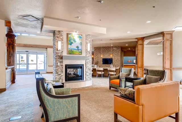 Communal lounge with armchairs and sofas arranged around a central double-sided fireplace, with a seating and reception area and entry doors visible in the background.