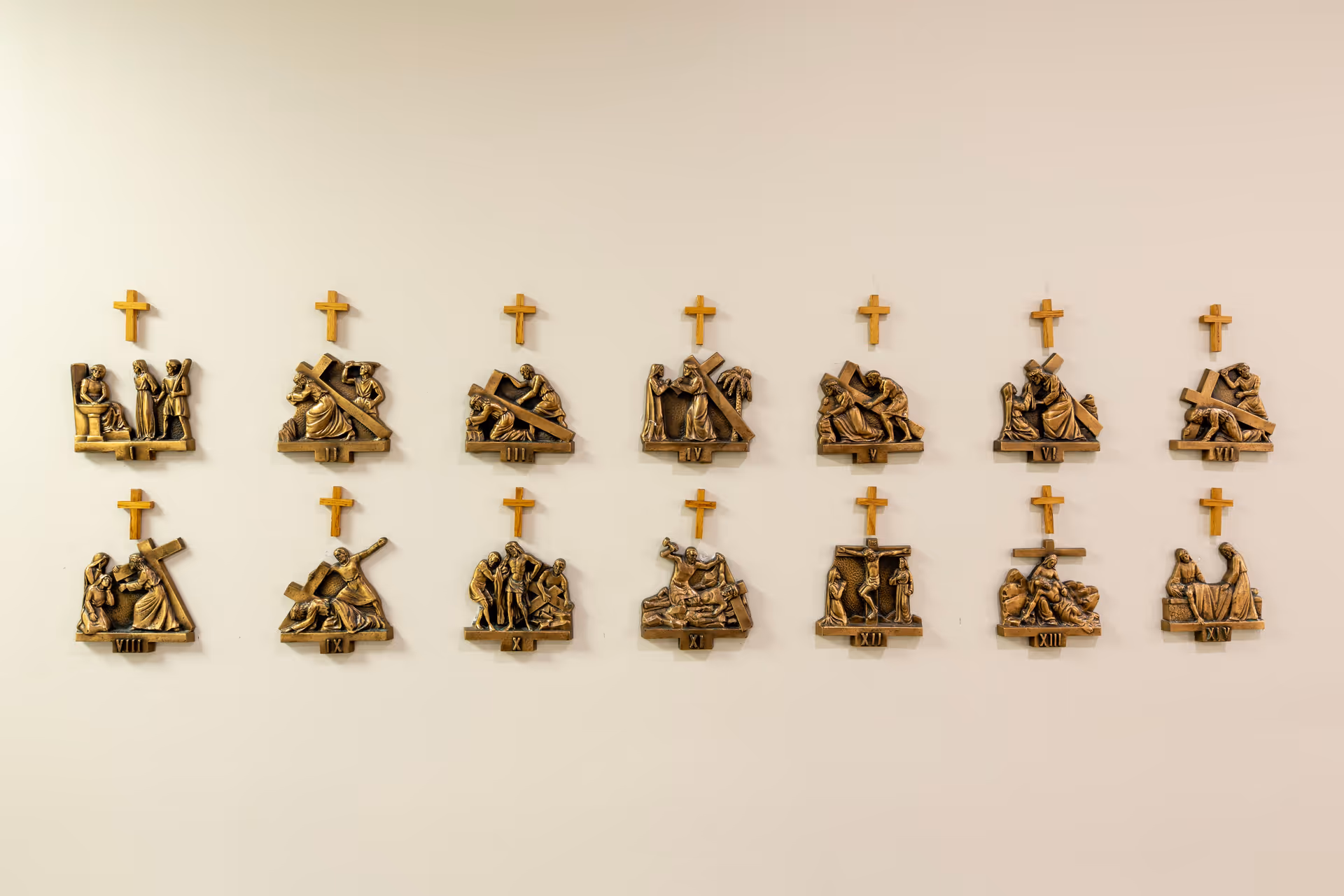 A series of fourteen bronze relief plaques mounted on a beige wall, each depicting a different scene from the Stations of the Cross, with small wooden crosses above each plaque.