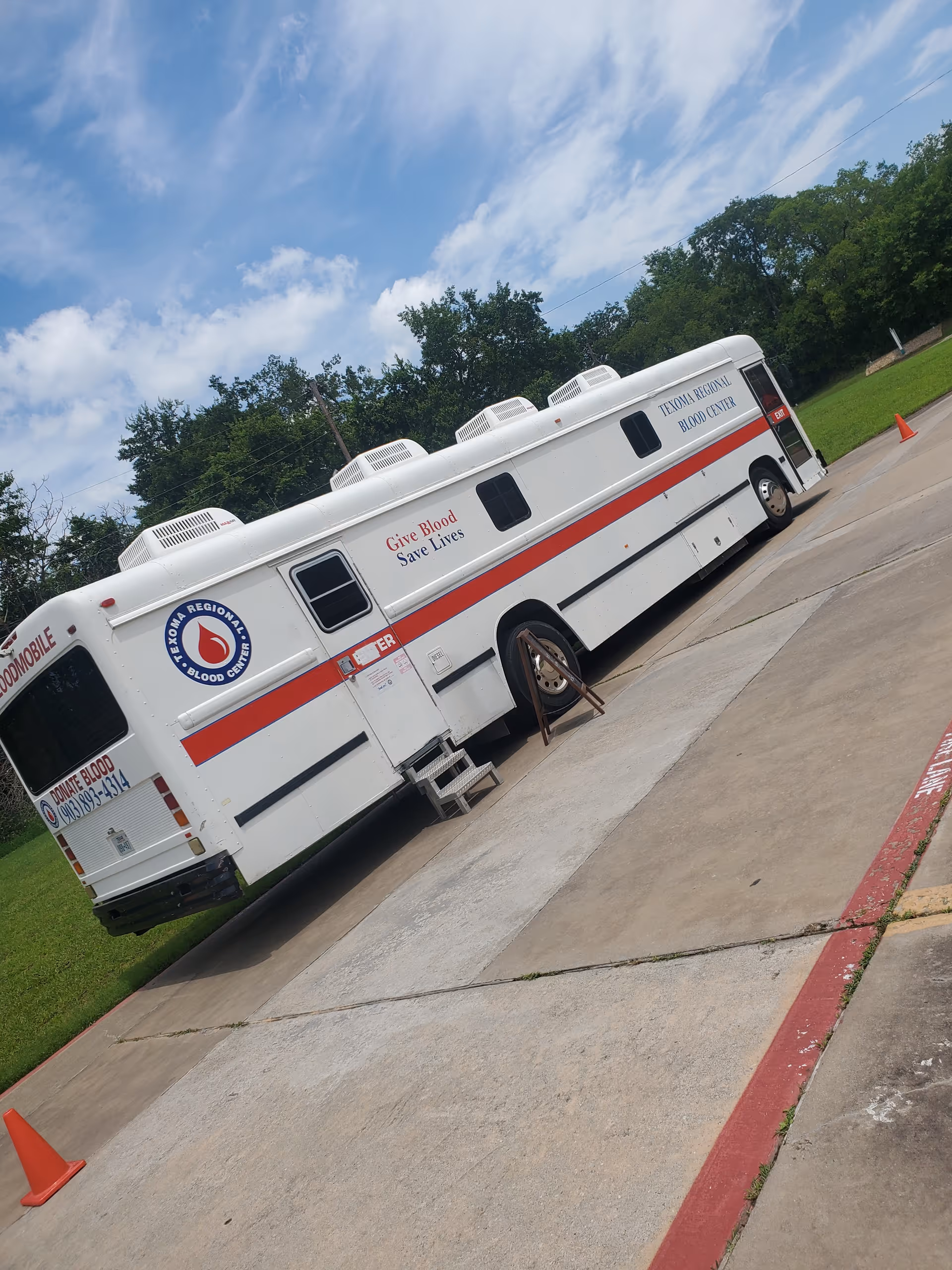 A white blood donation mobile unit with a red stripe parked on a driveway near trees.