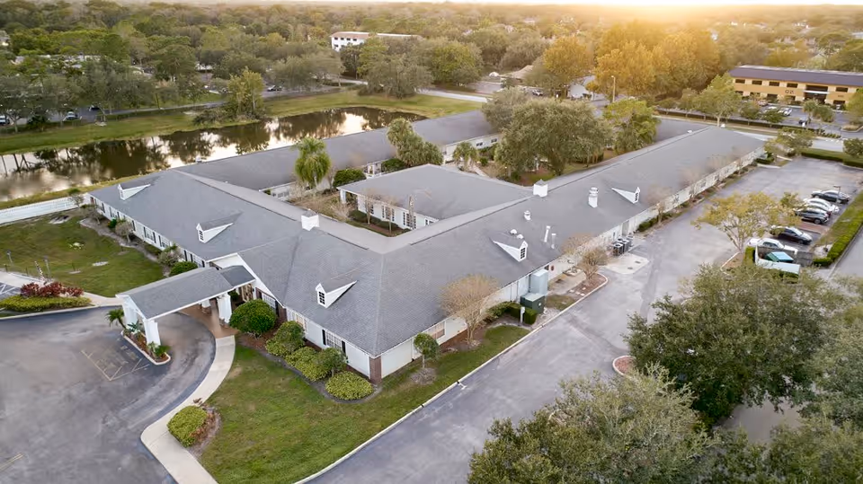 Aerial view of Palmetto Landing senior living facility showing a large, single-story building with a gray roof, surrounded by trees and greenery. There is a small pond nearby and a parking lot with several cars. The sun is setting in the background, casting a warm light over the scene.