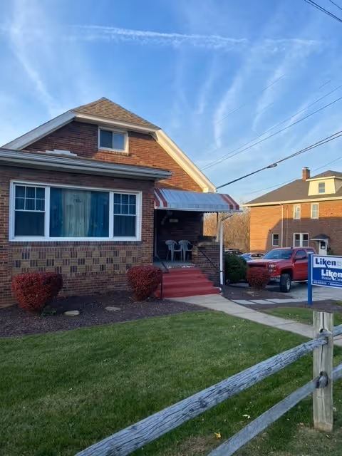 Front view of a brick house with awning, porch seating, and a Liken Home Care sign.