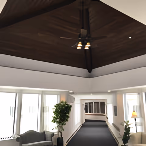 Interior view of a senior living facility hallway with a dark carpeted floor, white walls, large windows on the left side, and a high wooden ceiling with a ceiling fan and lights. There are green plants and seating areas along the hallway.