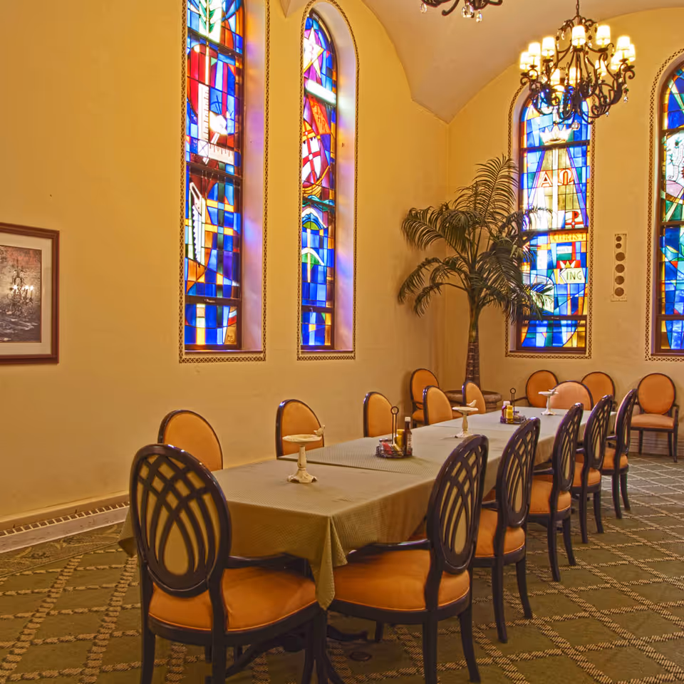 Long dining table with multiple chairs in a room featuring tall colorful stained-glass windows and a chandelier.