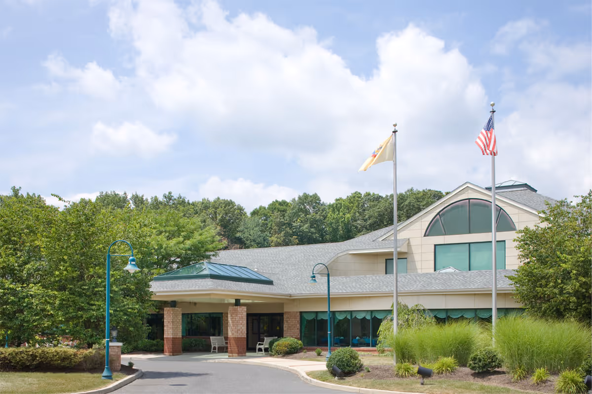 Front entrance of a senior living building with two flagpoles, a covered driveway, and landscaped greenery.
