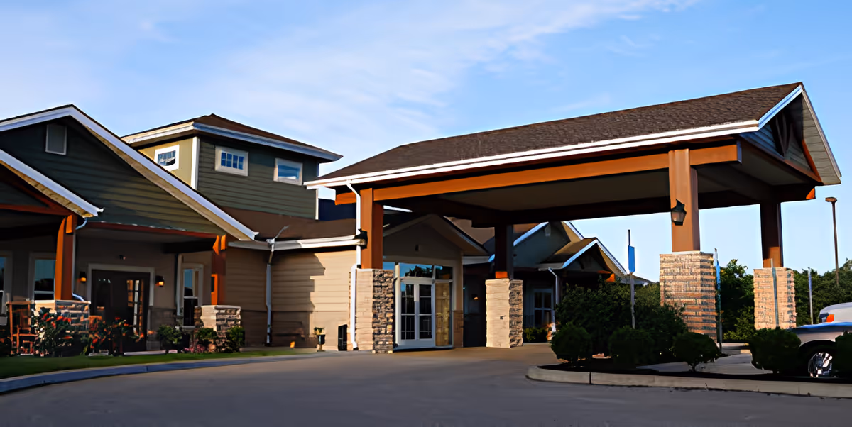 Covered porte-cochère and entrance of a senior living building with stone columns, landscaped shrubs, and a driveway.