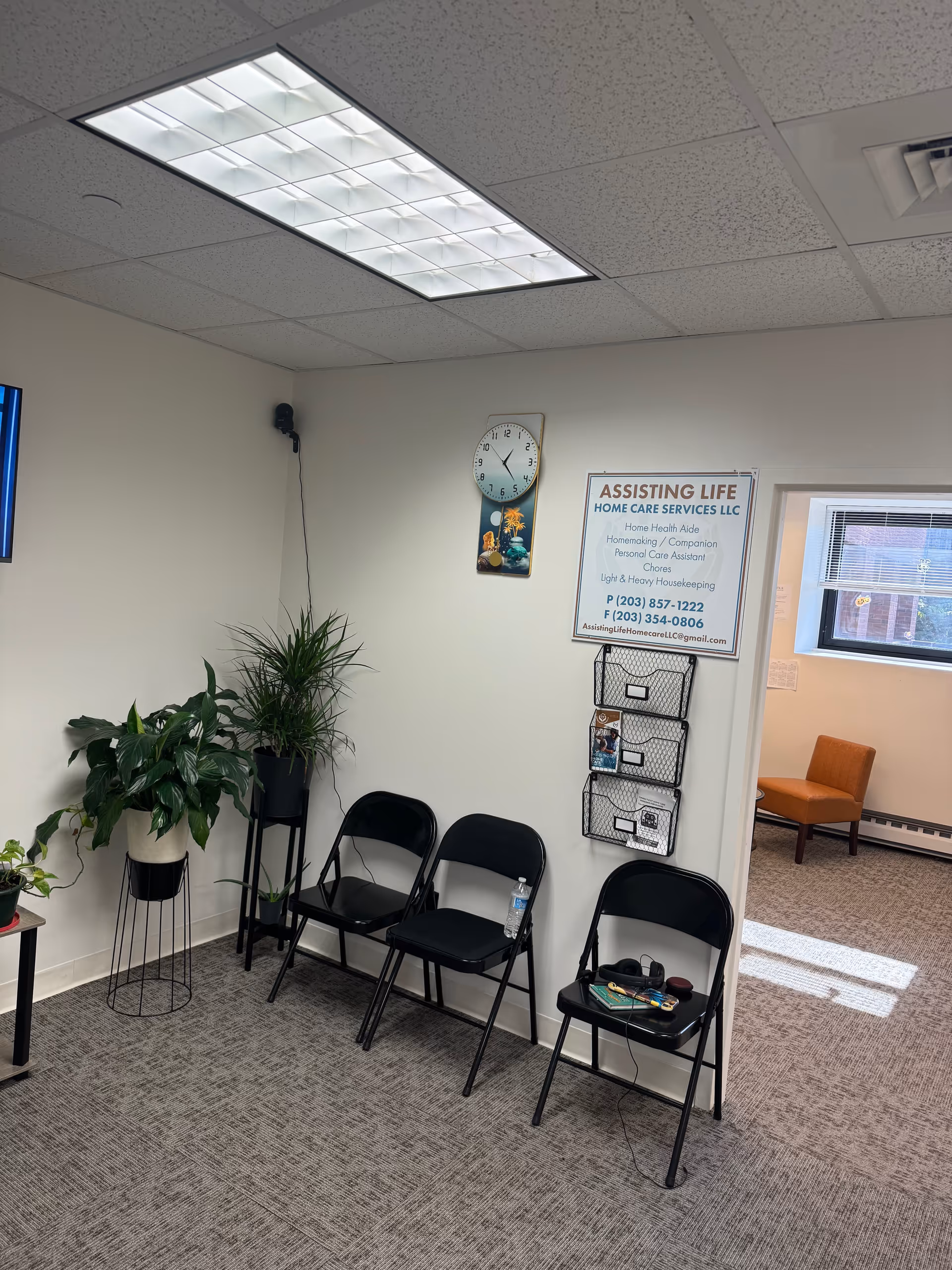 Interior waiting area with black chairs, plants, a wall clock, and a poster