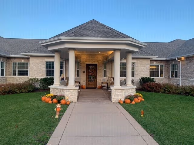 Front entrance of a single-story brick building with a covered porch supported by four columns. The walkway leading to the door is flanked by green grass and decorated with pumpkins and small scarecrows, suggesting a fall or Halloween theme. The sky is clear and blue.