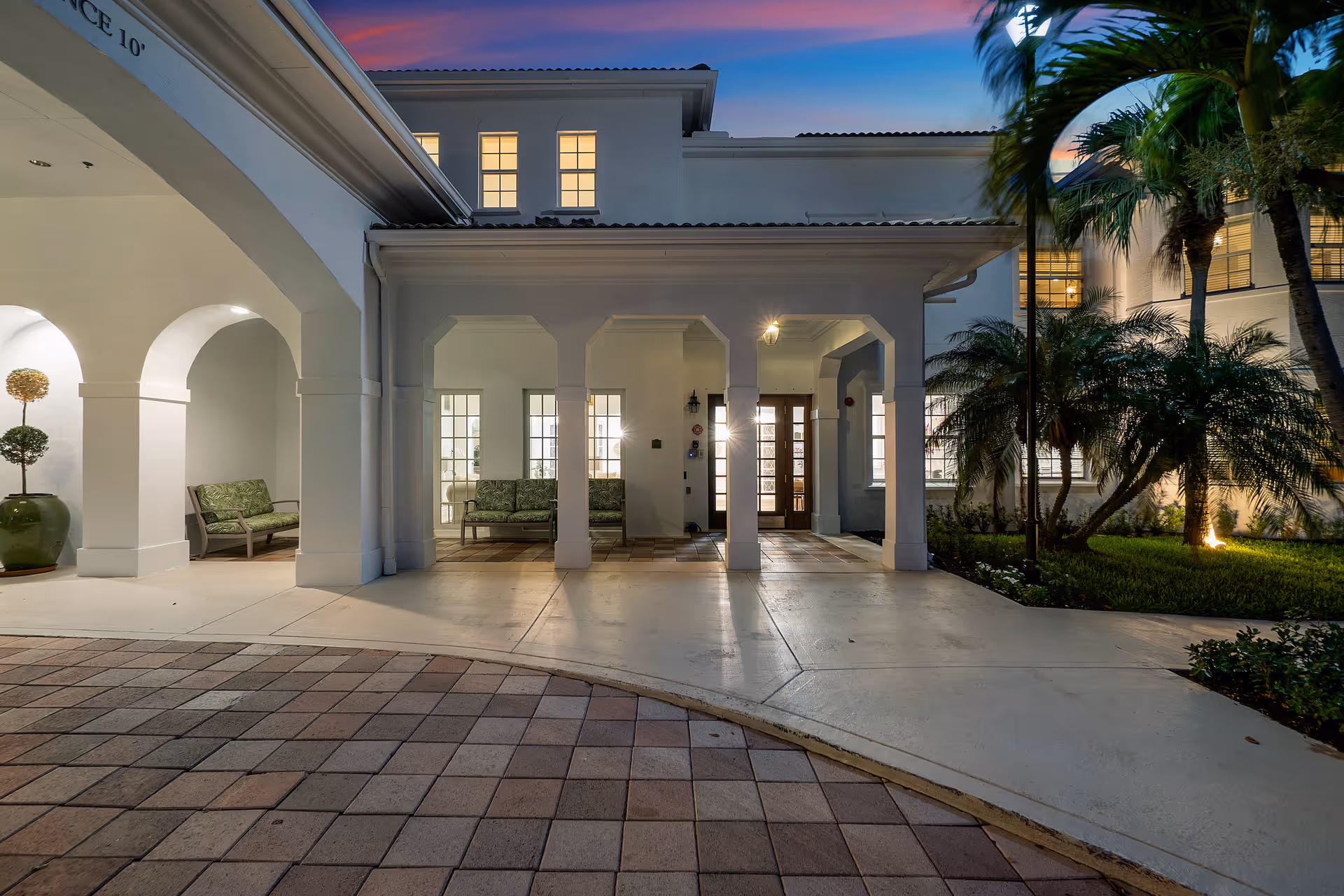 Exterior view of the entrance to a senior living facility named Tequesta Terrace at dusk. The building features white walls, arched columns, and large windows with warm lights inside. There are green cushioned benches under the covered entrance area and palm trees with landscaping on the right side.