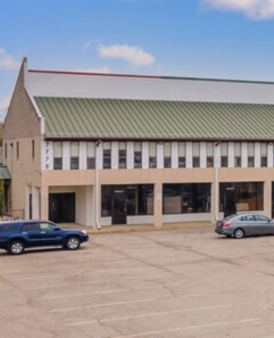 A front view of a beige two-story building with a green metal roof and parked cars.