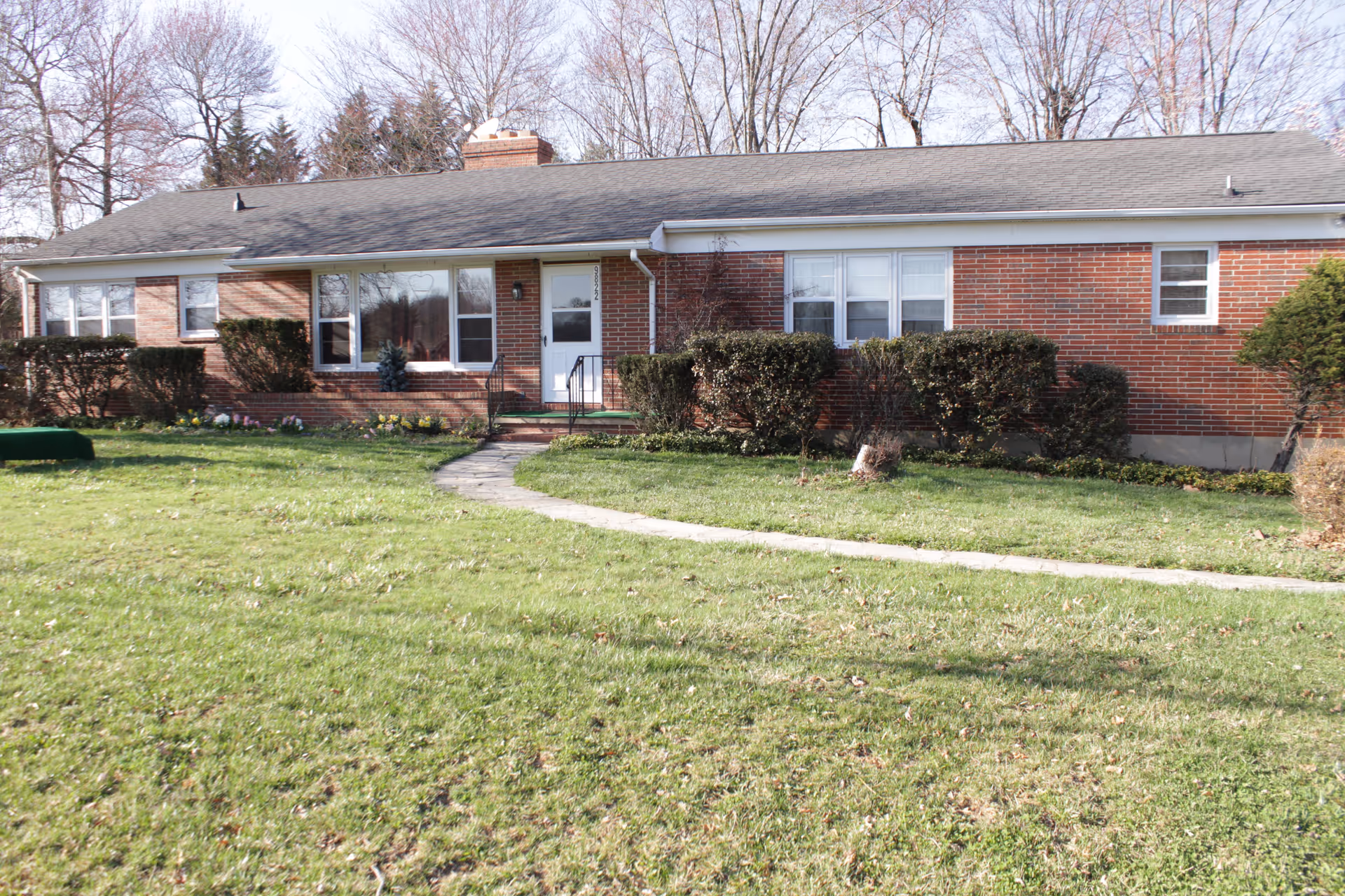 Single-story brick ranch-style house with a front lawn, walkway to the entrance, and trimmed bushes.