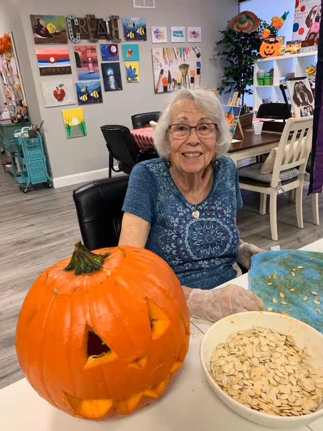An elderly woman with white hair and glasses is smiling while sitting at a table in a recreational room. She is wearing a blue patterned shirt and gloves, and in front of her is a carved pumpkin with a jack-o'-lantern face and a bowl filled with pumpkin seeds. The background shows colorful paintings on the wall and various decorations, including a Halloween-themed pumpkin decoration.