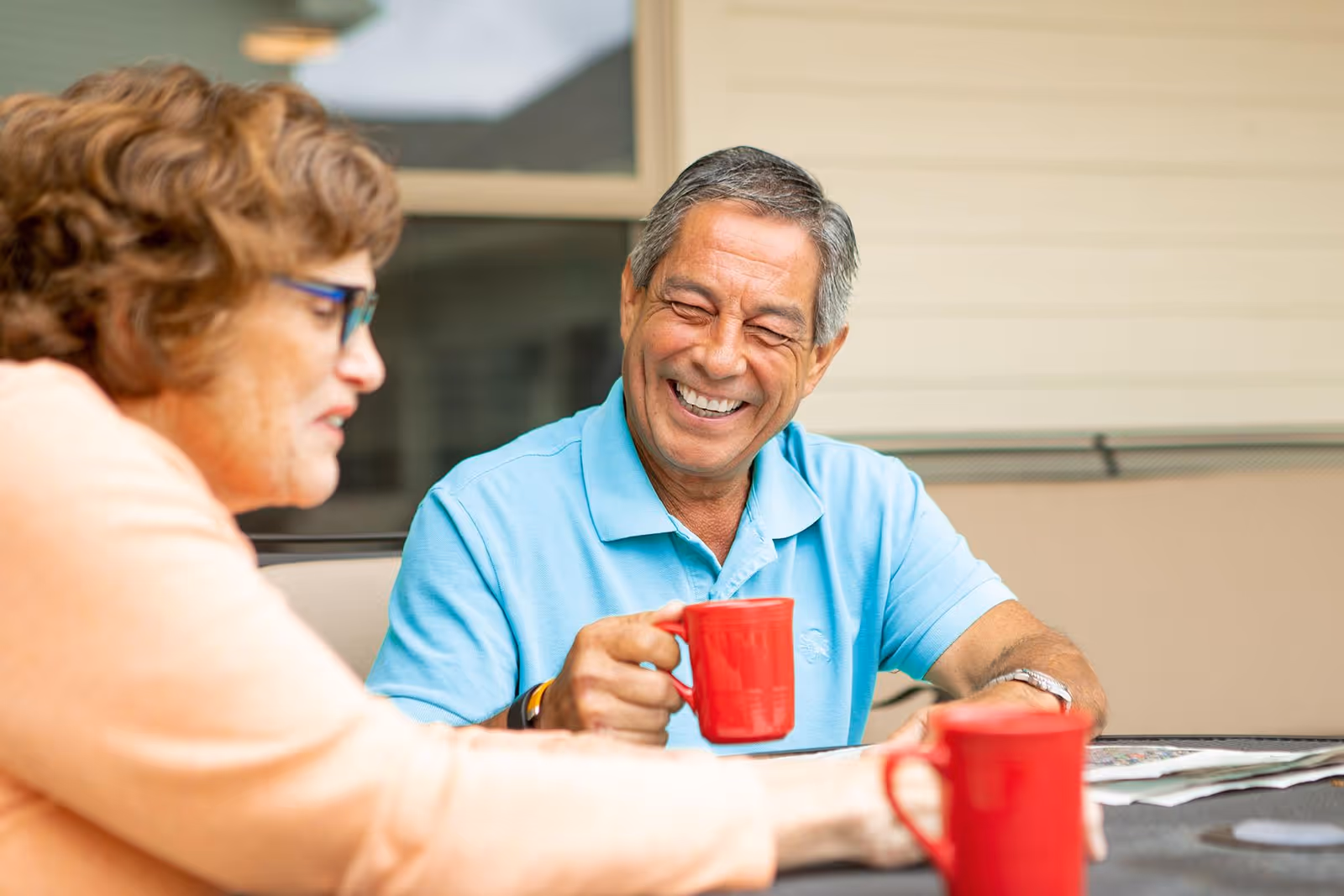Two elderly people sitting outdoors at a table, smiling and holding red mugs, with a beige building wall and window in the background.