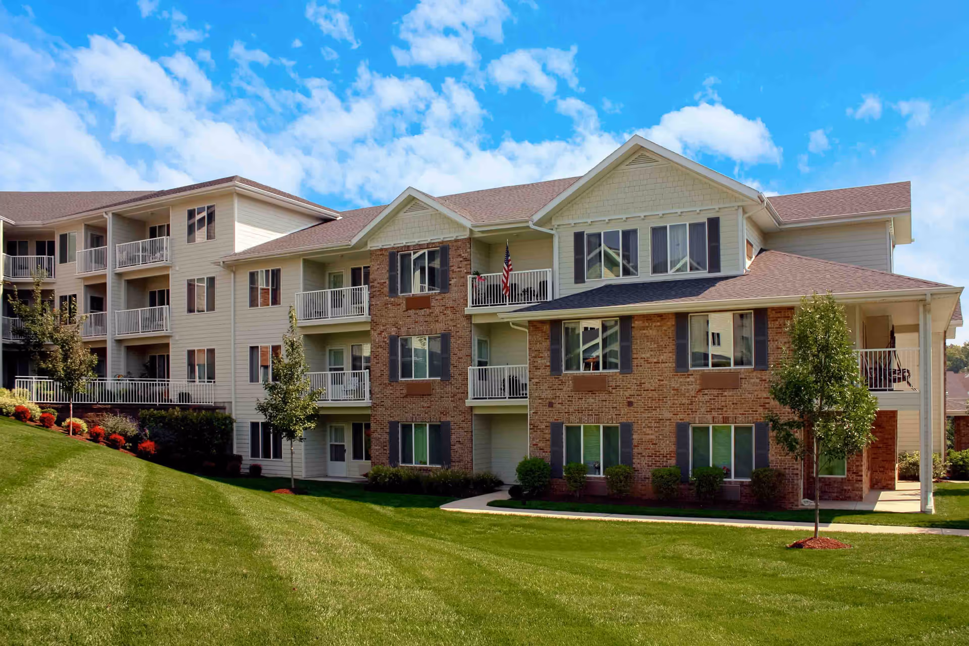 Three-story brick-and-siding senior living building with balconies, landscaped lawn, and a blue sky.