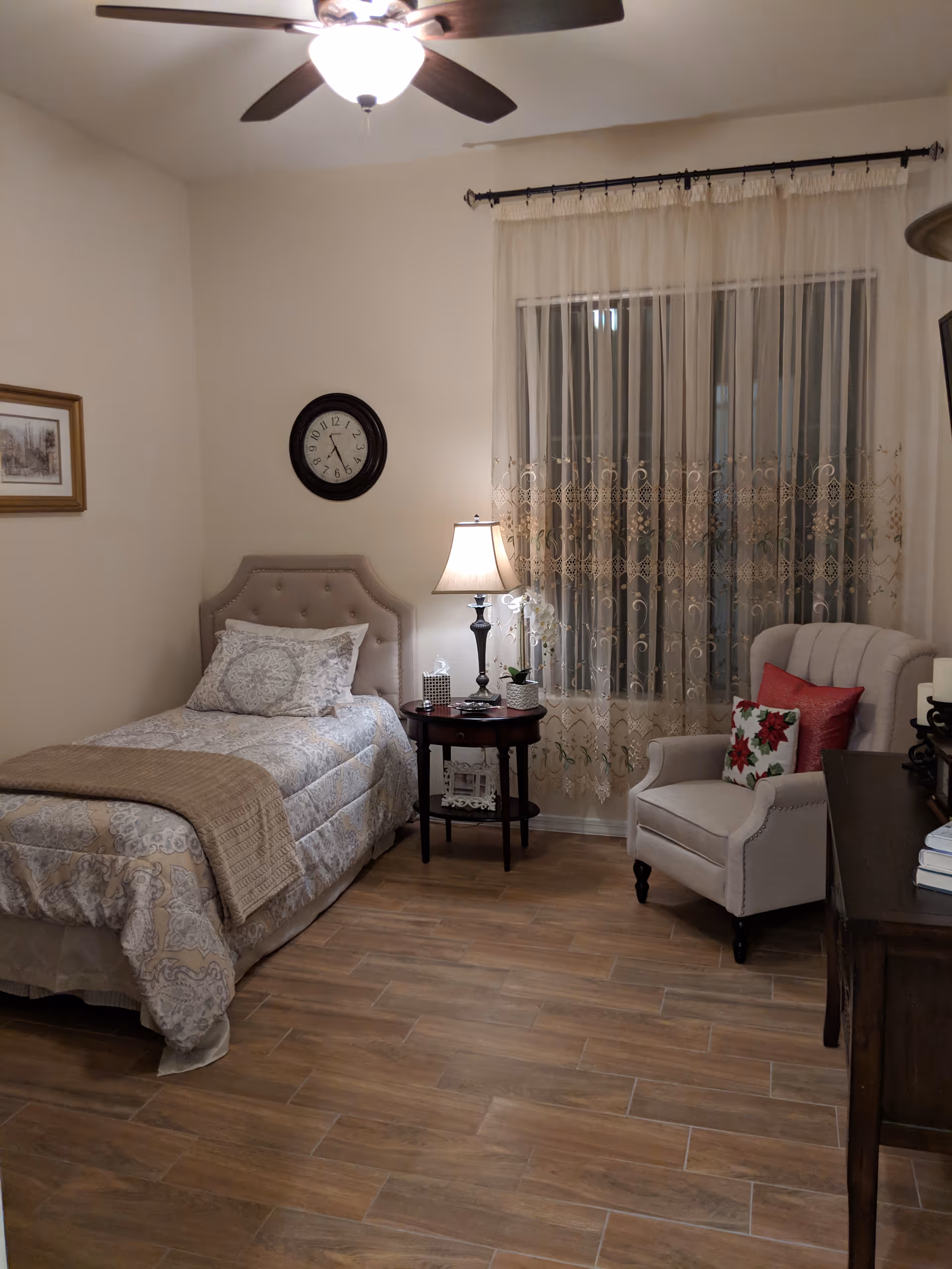 Cozy single bedroom with bed, nightstand, lamp, chair, and window with lace curtains.