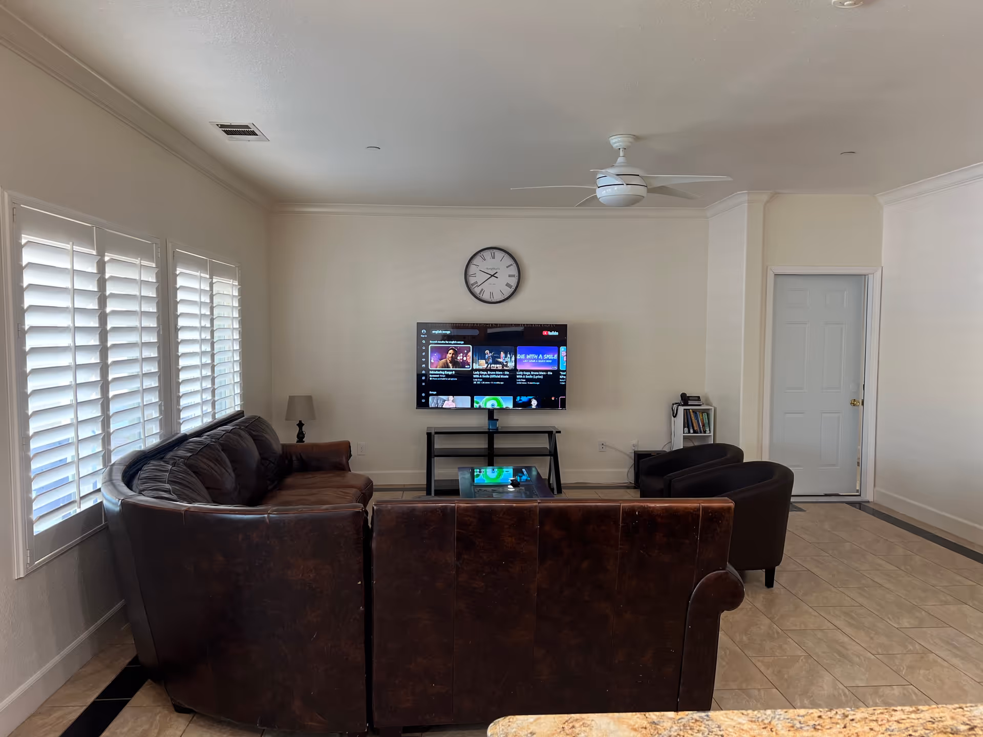 Senior care home living room with brown leather sofas, glass coffee table, TV, clock, and windows.