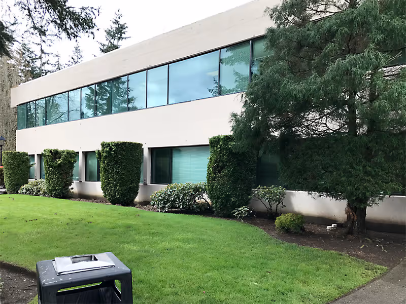 A modern beige building with large windows, trimmed shrubs, and a green lawn.