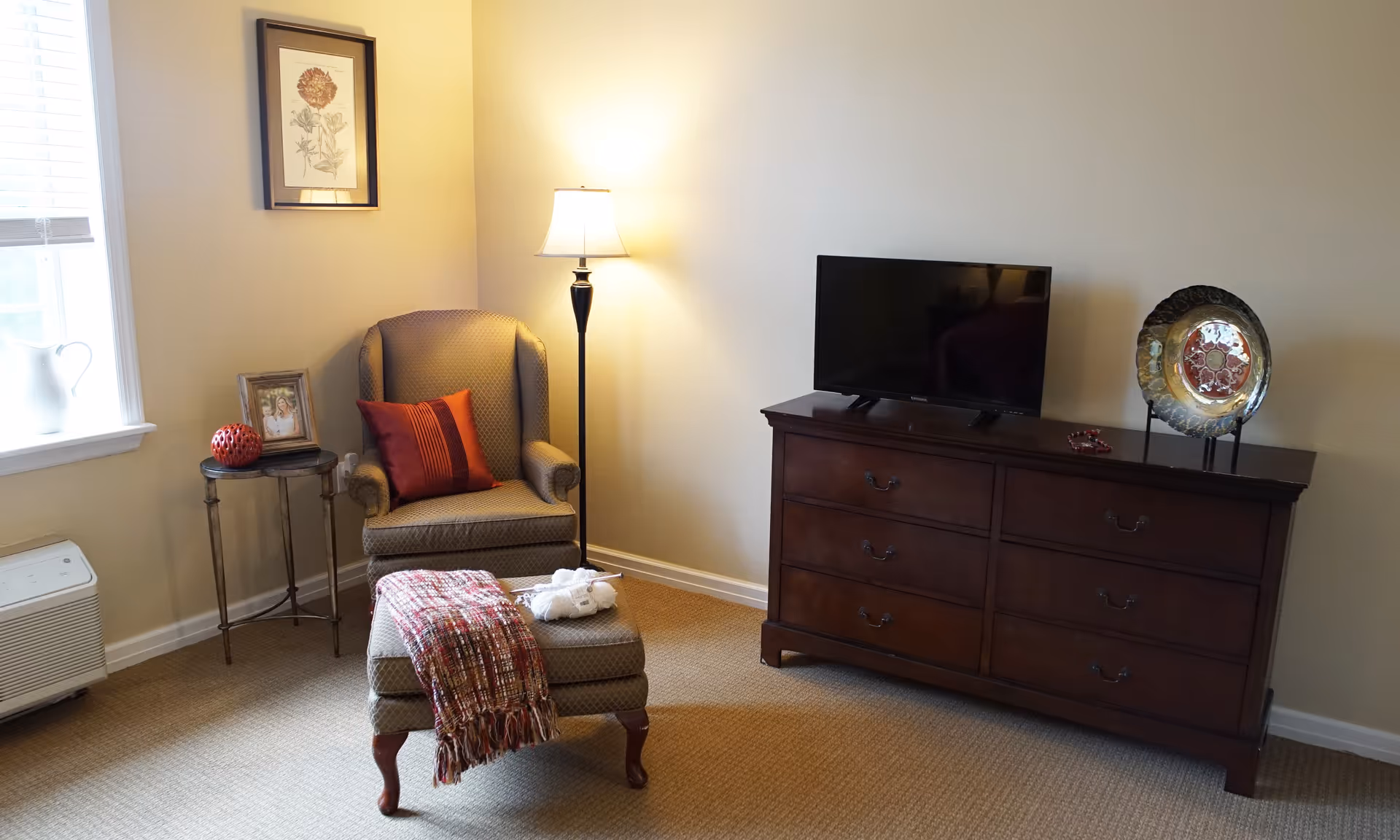 Cozy living room with armchair, ottoman, floor lamp, TV on a dresser, and a window.