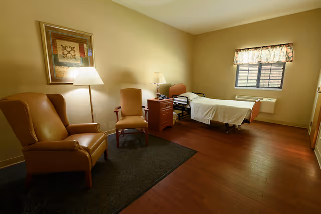 A simple senior care bedroom with a single hospital-style bed, nightstand and lamps, two chairs on a rug, framed artwork and a window.