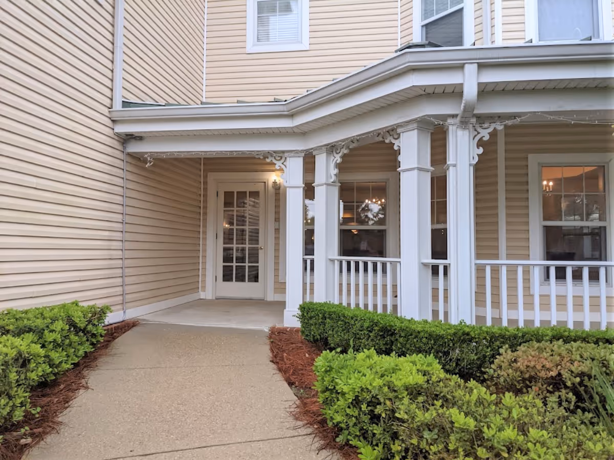 Covered entrance with white columns and railing leading to a glass-paneled door on a beige-sided building with trimmed shrubs.