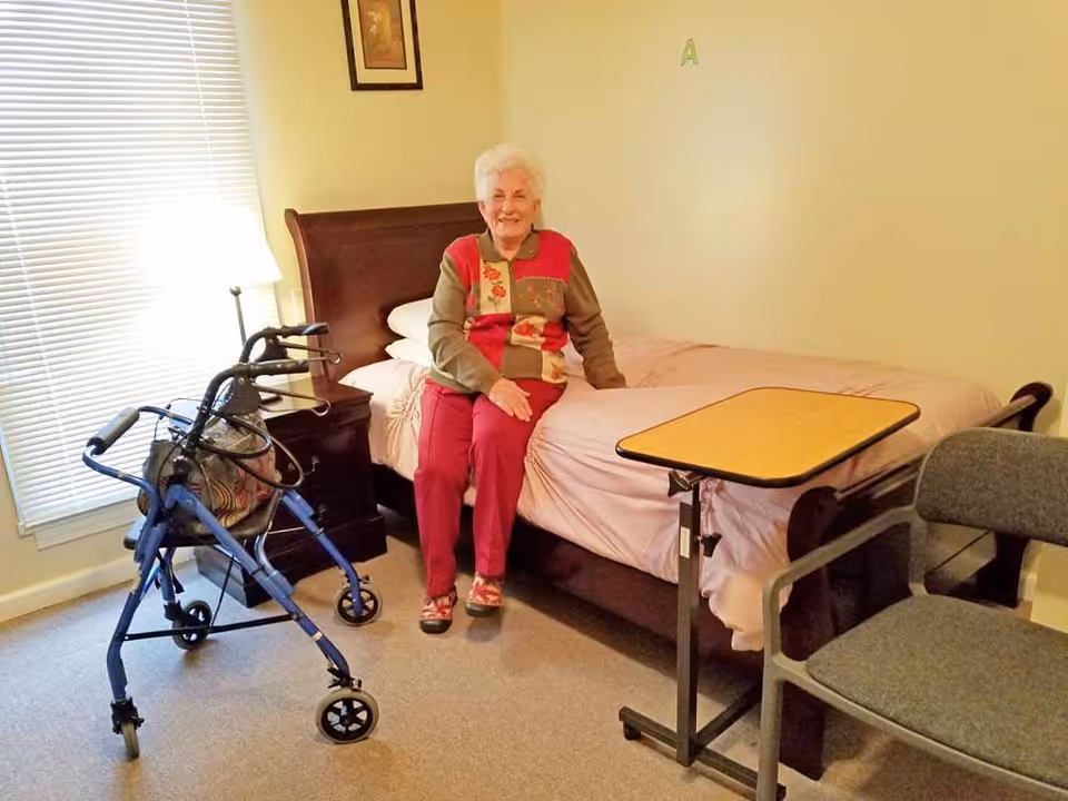 An elderly woman with white hair, wearing a colorful sweater and red pants, is sitting on the edge of a bed in a well-lit room. Next to her is a blue walker, a bedside table with a lamp, and a small overbed table. There is also a gray chair nearby. The room has beige walls and carpeted floor.
