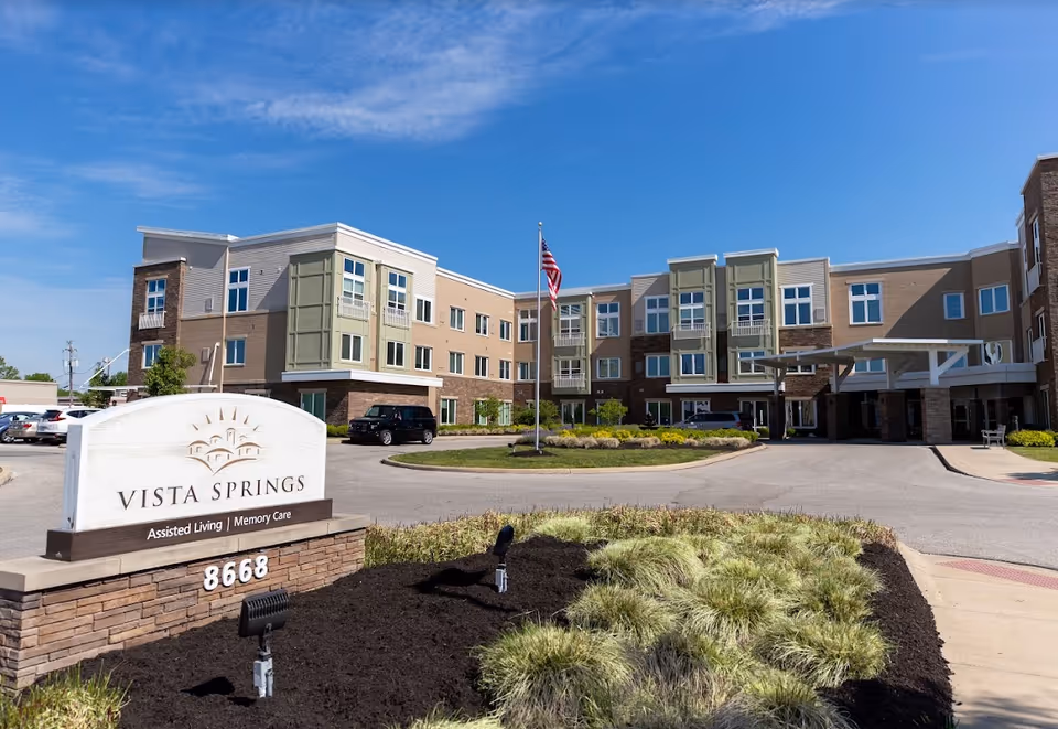 Exterior view of Vista Springs Greenbriar Village senior living facility on a clear sunny day, showing a three-story building with multiple windows, an American flag on a flagpole in front, a curved driveway, and a landscaped area with grass and plants. A large sign in the foreground reads 'Vista Springs Assisted Living | Memory Care 8668'.