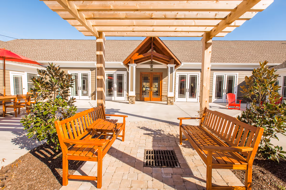 Sunlit courtyard with wooden benches under a pergola facing the entrance of a senior living building.