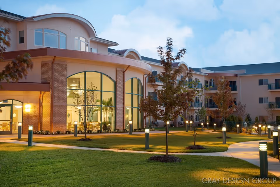 Exterior view of a senior living facility at dusk with well-lit pathways, manicured lawns, young trees, and large windows showing an interior dining area with tables and chairs.