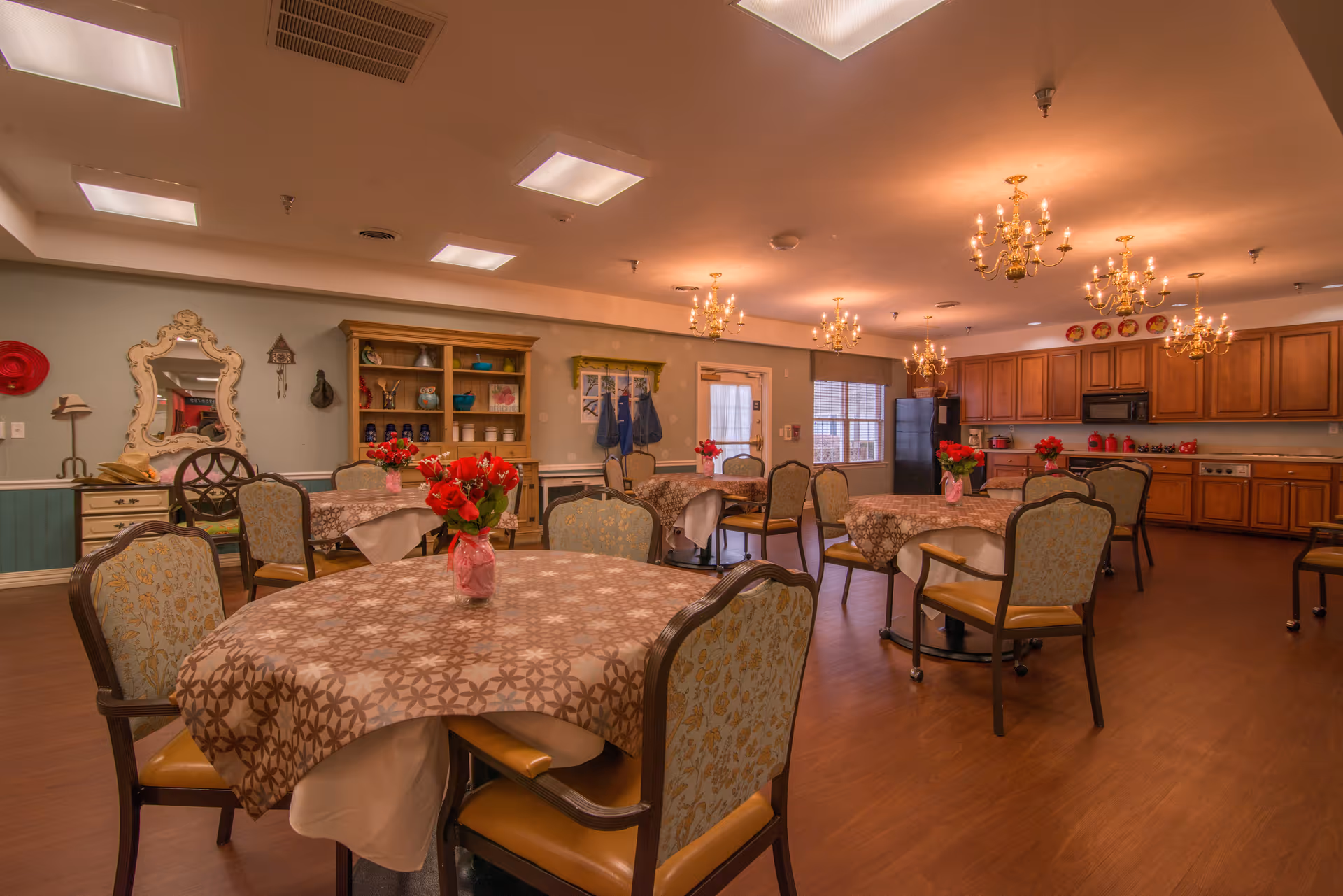 A dining room in a senior living facility with several round tables covered with patterned tablecloths and small vases of red flowers. The room has upholstered chairs with wooden frames, wooden cabinetry along one wall, a refrigerator, and multiple chandeliers hanging from the ceiling providing warm lighting. There is a decorative mirror, shelves with various items, and a window letting in natural light.
