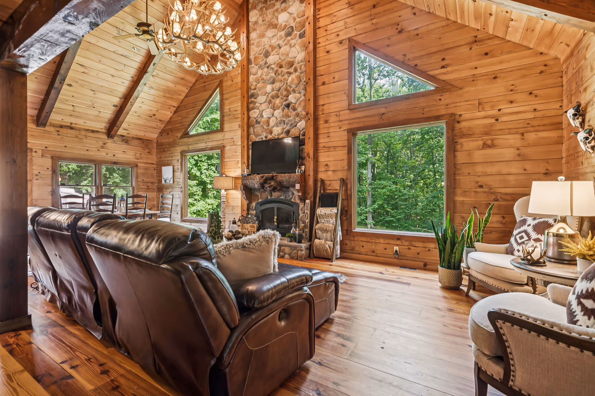 Rustic wooden living room with leather seating, stone fireplace, and large forest-view windows.