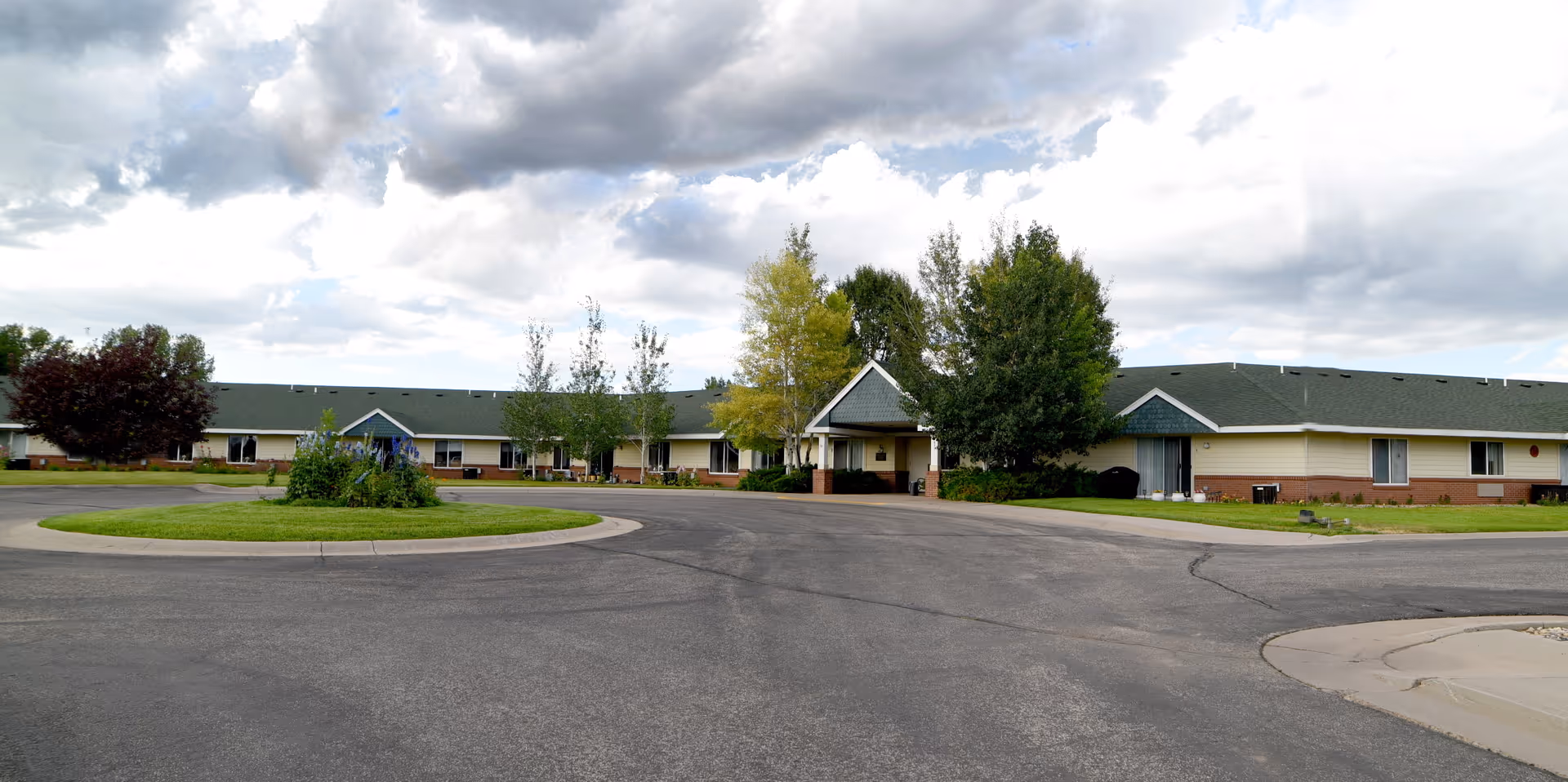 Single-story retirement residence building with a covered entrance, circular driveway and landscaped island under a cloudy sky.