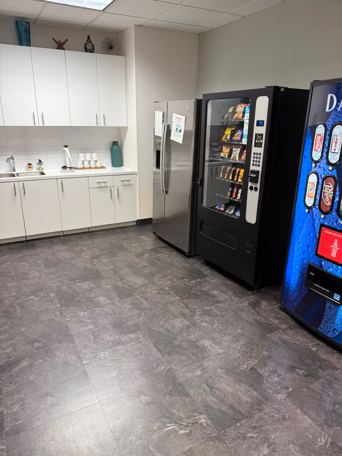 Industrial-style kitchenette with white cabinets, coffee station, sink, and vending machines.