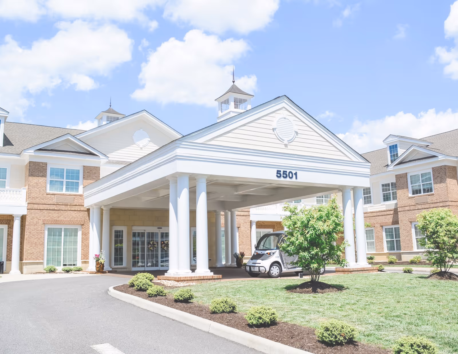 Front exterior view of a senior living facility named Edgeworth Park at New Town, featuring a covered entrance with white columns, a small vehicle parked under the entrance, well-maintained landscaping with bushes and trees, and a clear blue sky with some clouds.