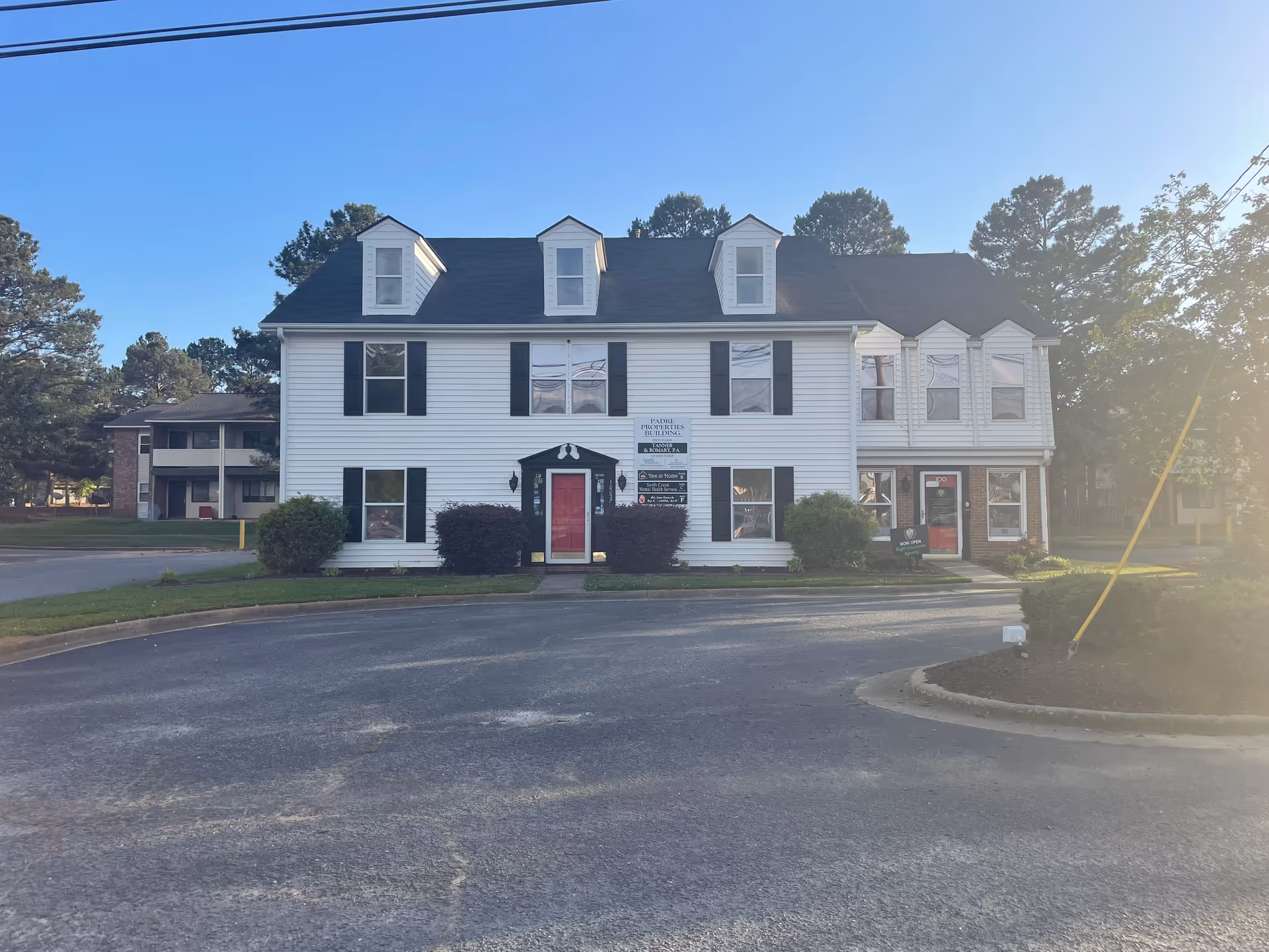 White two-story building with dormers and a red front door, parking lot in front.