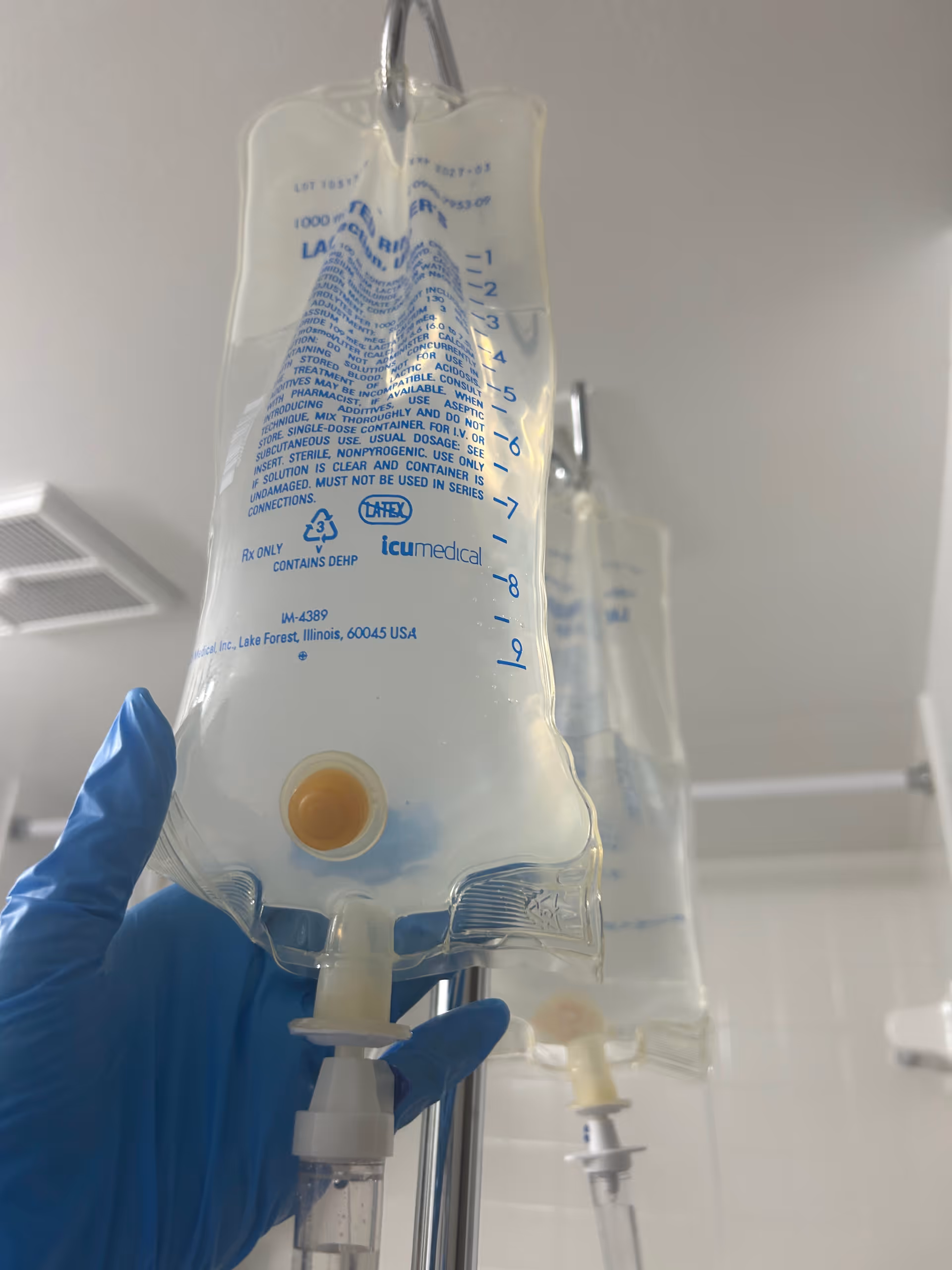 Close-up of an IV bag and tubing held by gloved hand in a clinical setting.