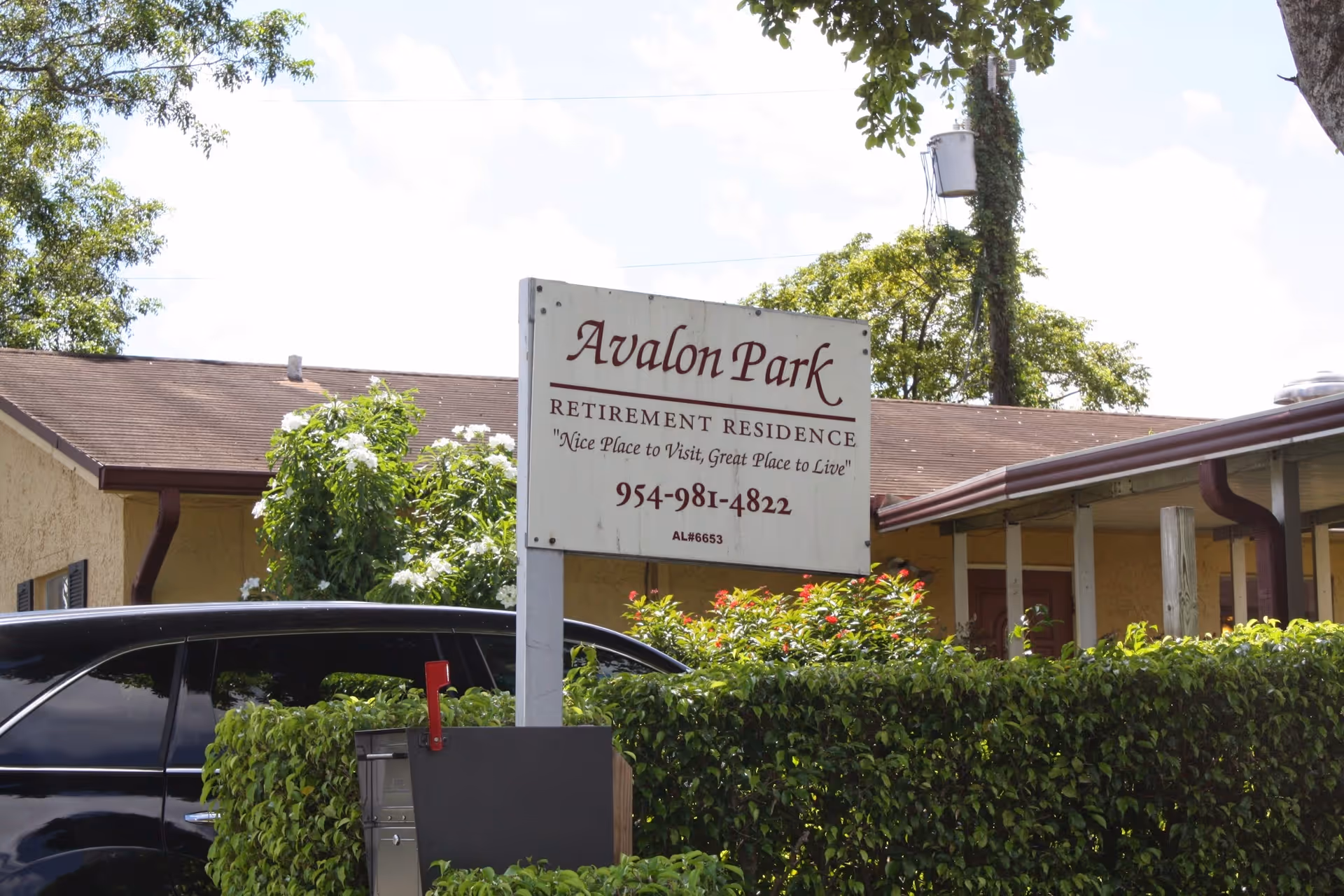 Outdoor view of Avalon Park Retirement Residence sign with the tagline 'Nice Place to Visit, Great Place to Live' and a contact phone number. The sign is surrounded by greenery and in front of a building with a brown roof and beige walls.