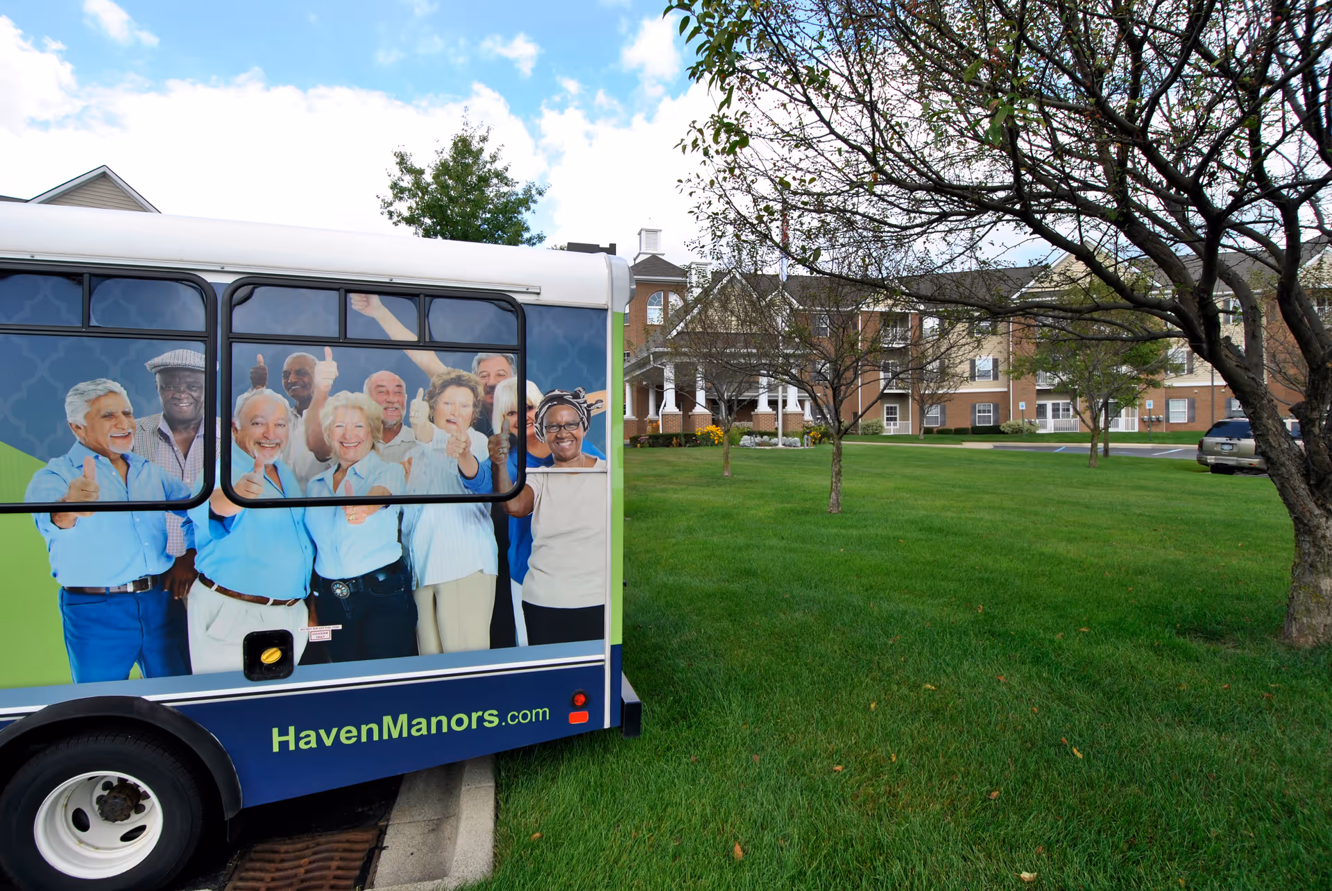A shuttle bus with a smiling seniors advertisement parked on a lawn in front of a brick senior living building.