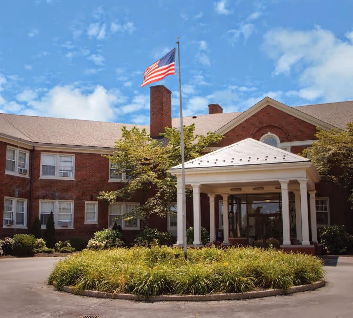 Front exterior view of Eliza Jennings Health Campus building with a circular driveway, an American flag on a flagpole, and greenery around the entrance under a partly cloudy blue sky.