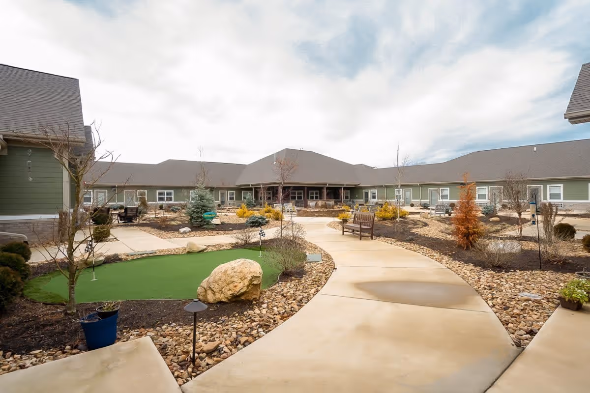 Outdoor courtyard area of a senior living facility with a curved concrete walkway, benches, small trees, shrubs, and a small artificial putting green. The building surrounds the courtyard with green siding and multiple windows under a cloudy sky.
