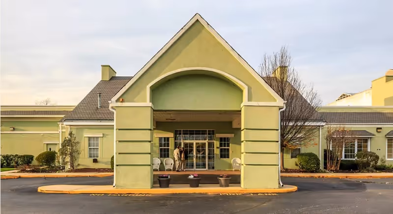 Front entrance of a pale green senior living facility with a covered portico and two people near the glass doors.