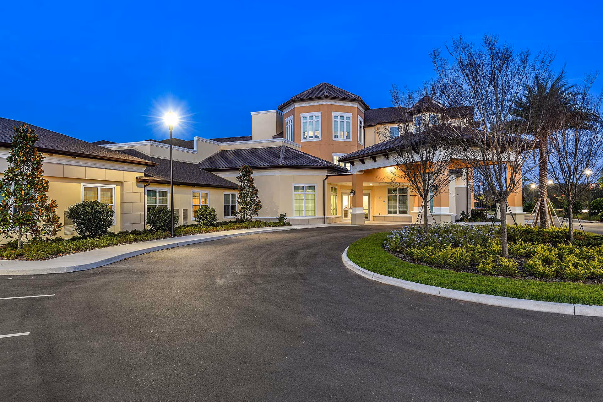Well-lit front entrance of a two-story senior living building with a curved driveway and landscaped island at dusk.