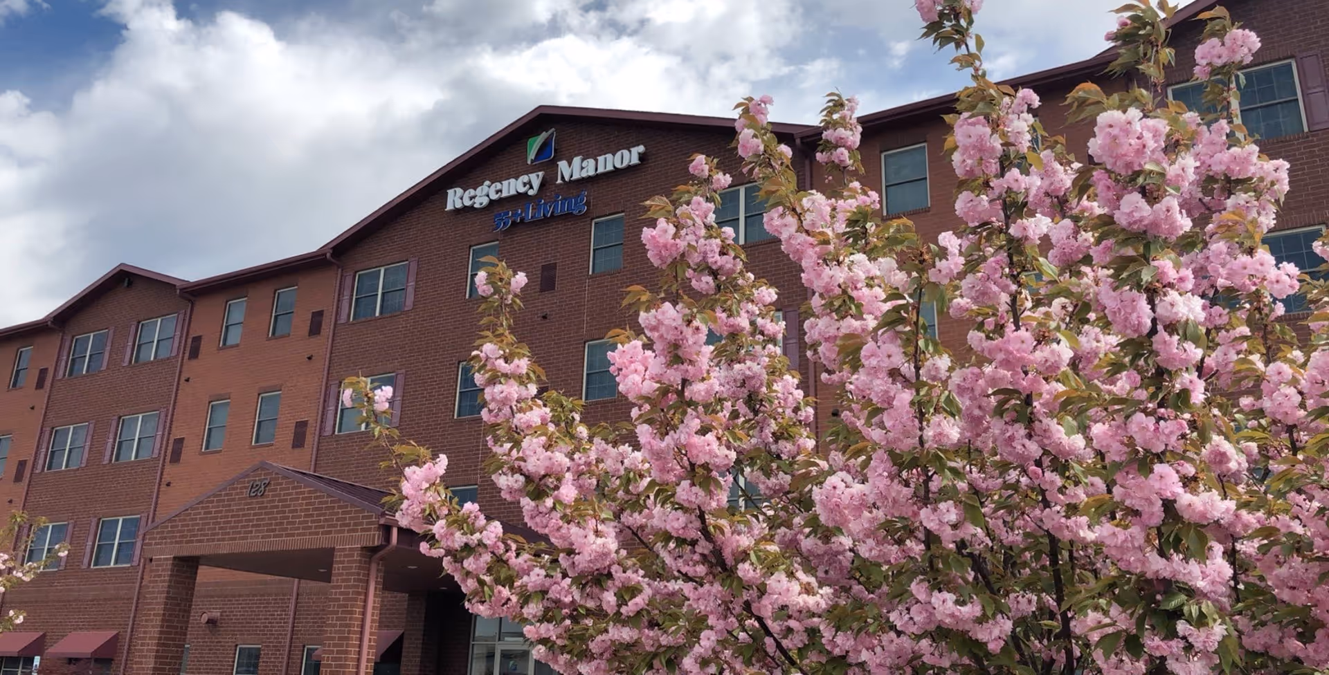 Exterior view of a multi-story brick building with the sign 'Regency Manor 55+ Living' on the top. In the foreground, there is a tree with abundant pink blossoms. The sky is partly cloudy.