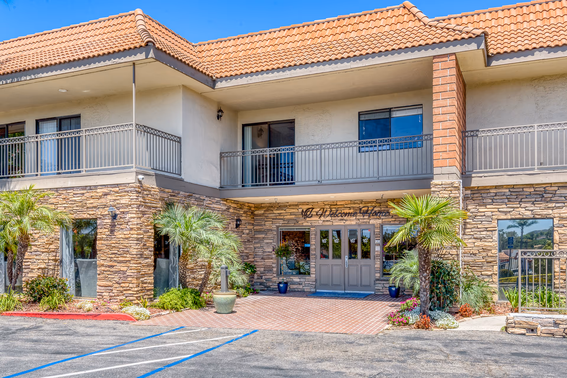 Exterior view of a senior living facility building with stone and stucco walls, a tiled roof, and a balcony. The entrance has double doors with glass panels and a sign above that reads 'Welcome Home'. There are palm trees and various plants around the entrance, and a parking area with marked spaces in front.