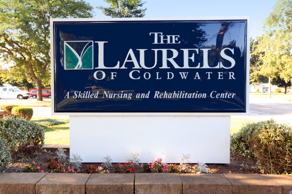 Outdoor sign for The Laurels of Coldwater, a skilled nursing and rehabilitation center, surrounded by bushes and flowers with trees and parked cars in the background.